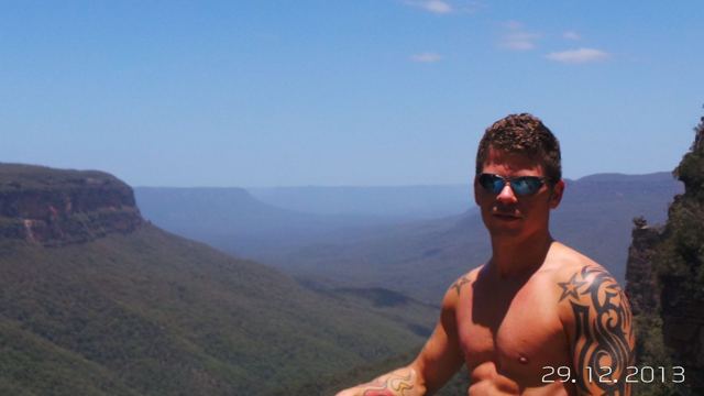 A man in sunglasses at a cliff-edge lookout over the forested valleys of the Blue Mountains.