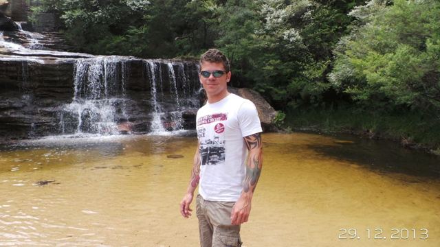 A man standing in a shallow golden pool below a small waterfall in the Blue Mountains.