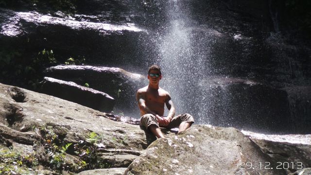 A man sitting on a rock beneath a waterfall in a dark gorge.
