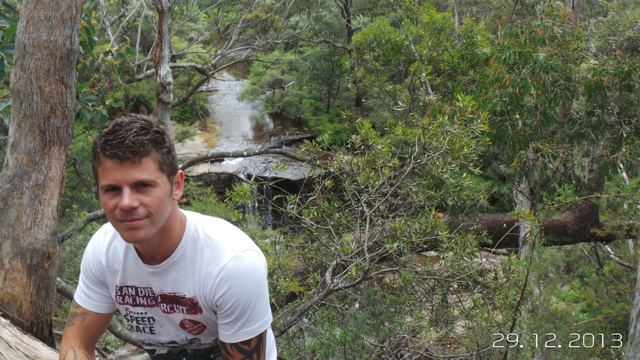 A smiling man in a white T-shirt in front of a small waterfall in the bush.