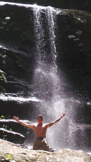 A man raising his arms beneath a tall waterfall cascading down a dark cliff.