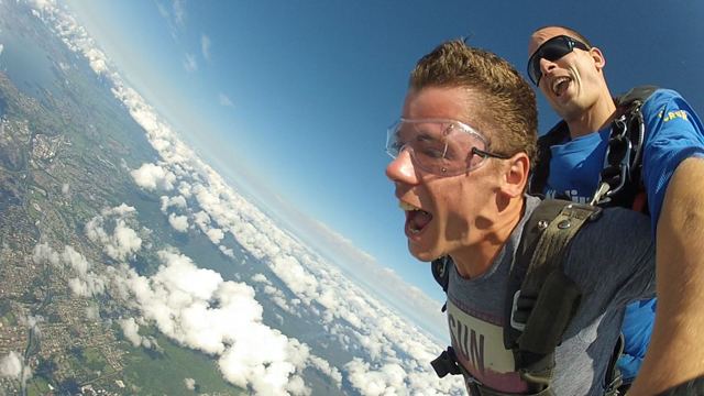 A man in goggles shouting during a tandem skydive with clouds and coastline far below.