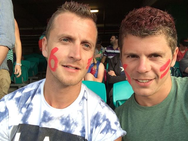 Two men with red numbers painted on their cheeks taking a selfie in a cricket stadium.