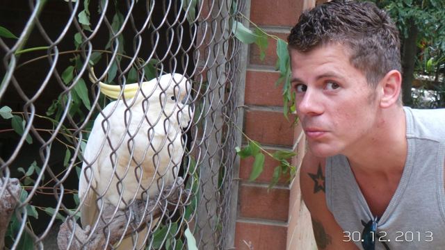 A man pulling a face next to a white cockatoo behind a wire fence.
