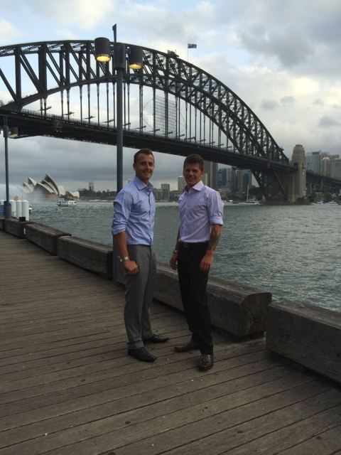 Two men in shirts standing on a wooden wharf with the Sydney Harbour Bridge and Opera House behind.