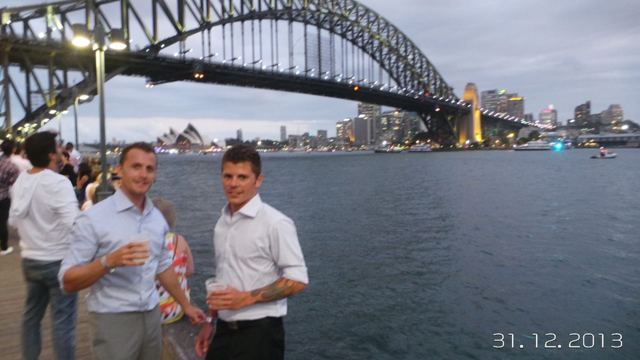 Two men holding drinks by the harbour with the Sydney Harbour Bridge and Opera House behind them.