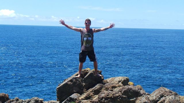 A man with arms outstretched standing on rocks above a deep blue sea.