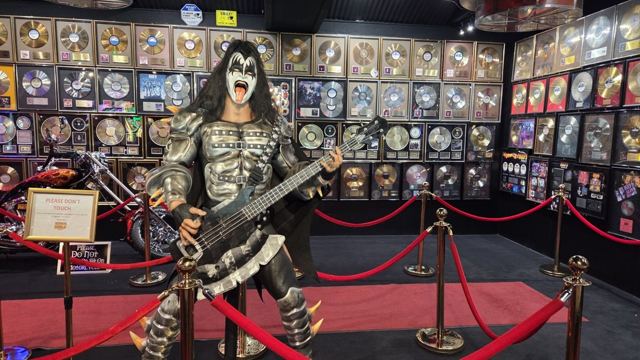 Life-size statue of a face-painted rock musician with a bass guitar in front of a wall of framed gold records.