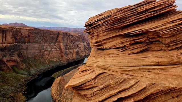 Layered, wave-like orange sandstone rock at the edge of a deep canyon with a river below.