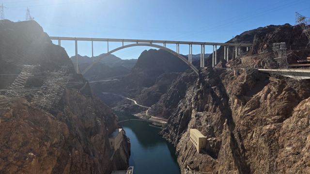 A concrete arch bridge spanning a deep rocky canyon with a river at the bottom, sun shining from above.