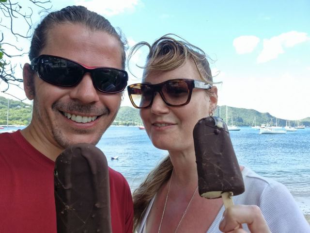 A man and a woman in sunglasses holding chocolate ice lollies by a harbour.