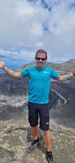 A man with arms out standing on the edge of a huge volcanic crater.