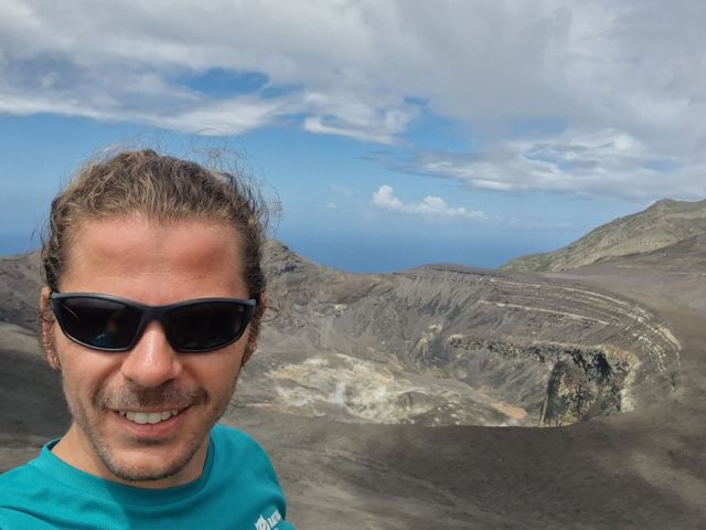 A man in sunglasses taking a selfie at the rim of a vast grey volcanic crater.