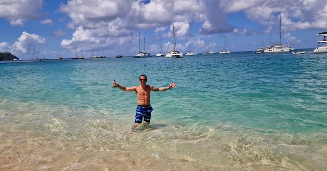 A man standing waist-deep in clear turquoise water with arms outstretched and yachts anchored behind.