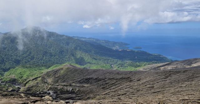 A view from a volcano's ash-covered slopes over green hills to the sea under a cloudy sky.