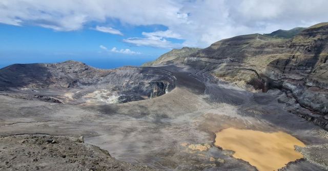 A vast grey volcanic crater with steep walls and a pale sandy patch on its floor.