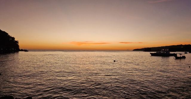 An orange sunset over calm sea with silhouetted boats and a dark headland.