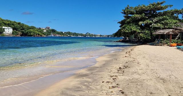 A golden sandy beach curving round to a tree-covered headland beside clear blue water.