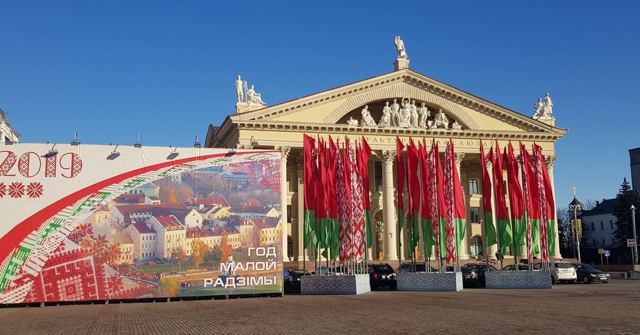 A grand neoclassical building fronted by rows of red and green Belarusian flags and a large mural.