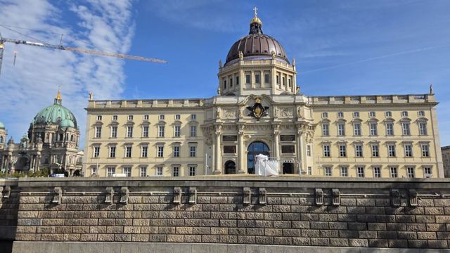 A large classical palace building with a domed roof next to a cathedral dome under a blue sky.