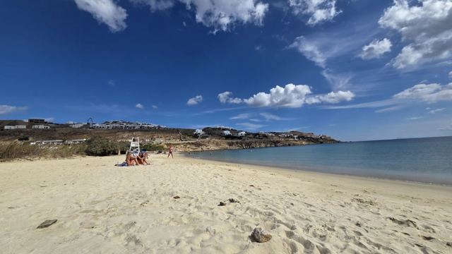 A wide sandy beach on Mykonos under a deep blue sky with scattered white clouds and calm sea.