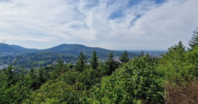 A view over dense forest and rolling hills under a cloudy blue sky.