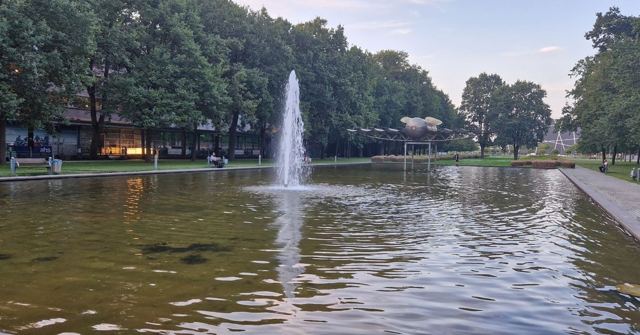 A tall fountain in a long rectangular pool surrounded by trees in a city park.