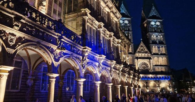 Illuminated stone arches of Bremen town hall with the cathedral towers lit up behind at night.