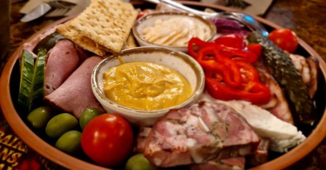 A round platter of cured meats, cheese, red peppers, olives, tomato and mustard.
