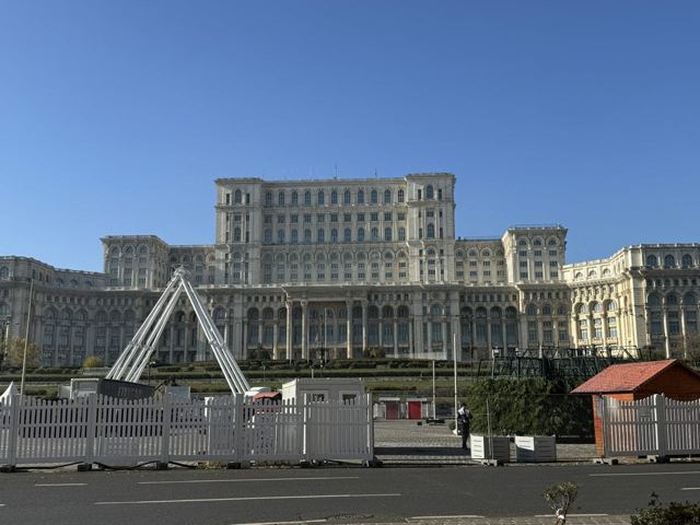 Enormous white neoclassical government building behind a road and fencing under a blue sky.