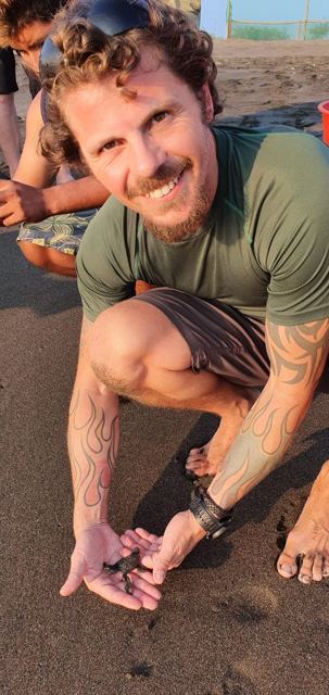A smiling bearded man crouching on black sand.