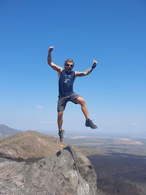A man jumping in the air on a rocky volcanic summit above a wide landscape.