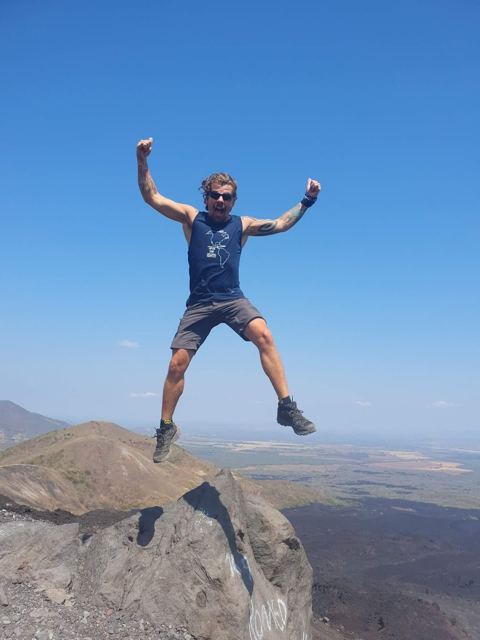 A man leaping with arms raised on a volcanic peak against a blue sky.
