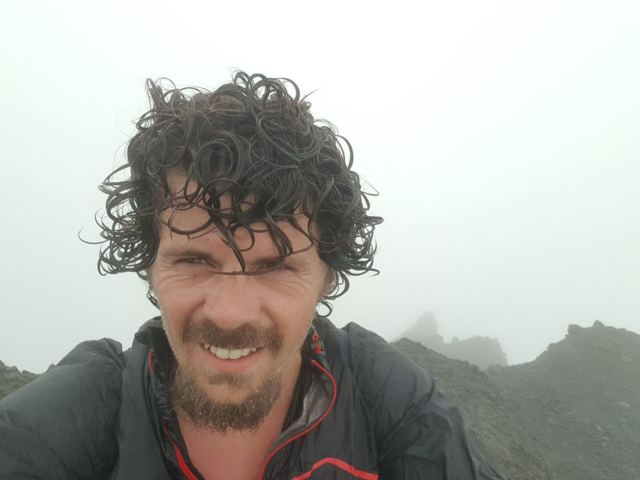 A smiling man with curly windswept hair taking a selfie in the mist on a volcano summit.