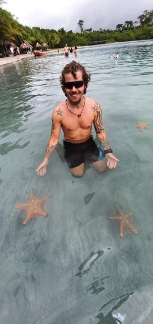 A shirtless man kneeling in clear shallow water surrounded by orange starfish.