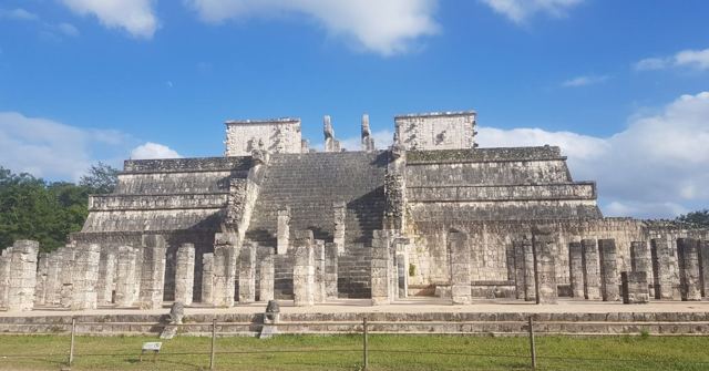 A stepped Mayan stone temple fronted by rows of columns on green grass under blue sky.
