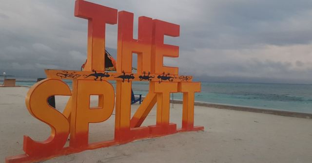 Big orange letters reading The Split on a pale beach beside the sea under grey sky.