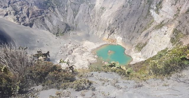 A turquoise lake at the bottom of a grey volcanic crater with scrubby vegetation.