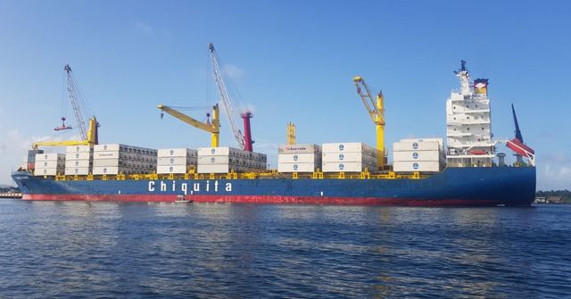 A large blue cargo ship with cranes and containers on calm water under blue sky.