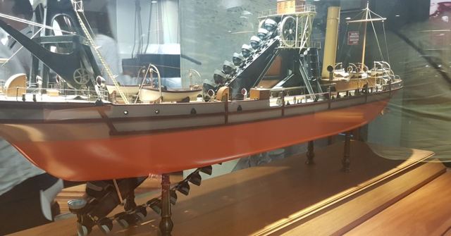 A detailed model ship with dredging machinery displayed in a glass case.