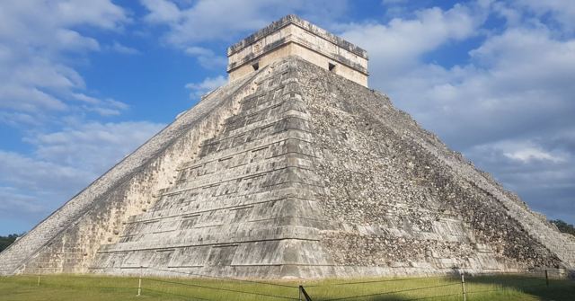 The stepped pyramid of El Castillo at Chichen Itza under a bright blue sky with clouds.