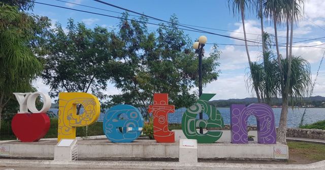 A colourful Peten sign beside a lake with palm trees and greenery.