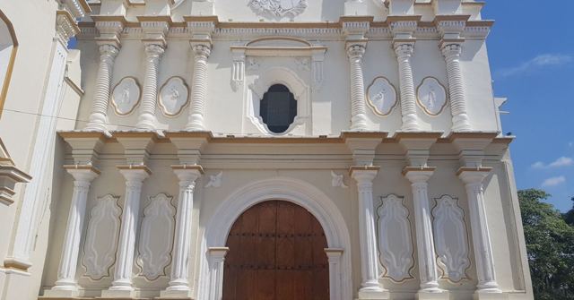 Ornate white baroque church facade with a large arched wooden door under blue sky.
