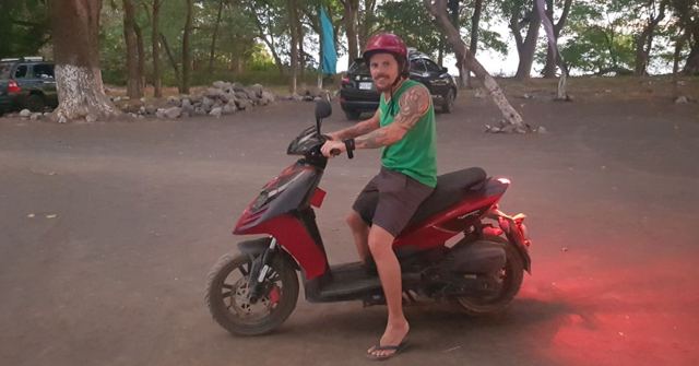 A man in a green vest and red helmet sitting on a red scooter on a dusty road.