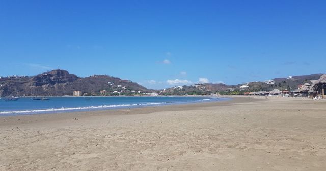 A wide sandy beach curving around a blue bay backed by green hills.