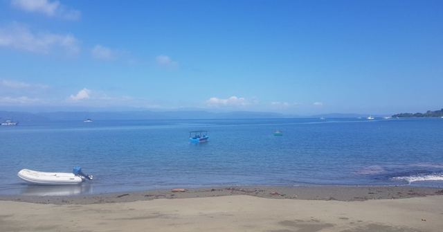 A calm blue bay with a small dinghy on a dark sandy beach and boats on the water.