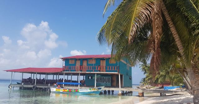 A colourful wooden house on stilts over the water with small boats and palm trees.