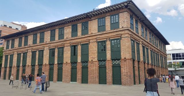 A large red-brick building with many green shuttered windows on a sunny plaza.