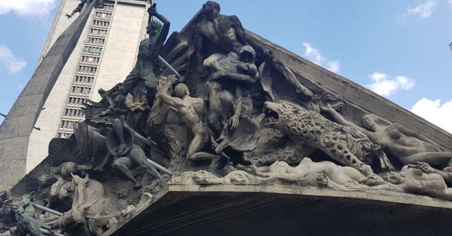 A large dark sculpture of intertwined figures rising diagonally against a tall building and blue sky.