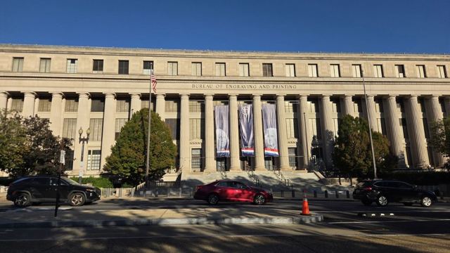 A long neoclassical stone government building with columns and hanging banners, trees in front.
